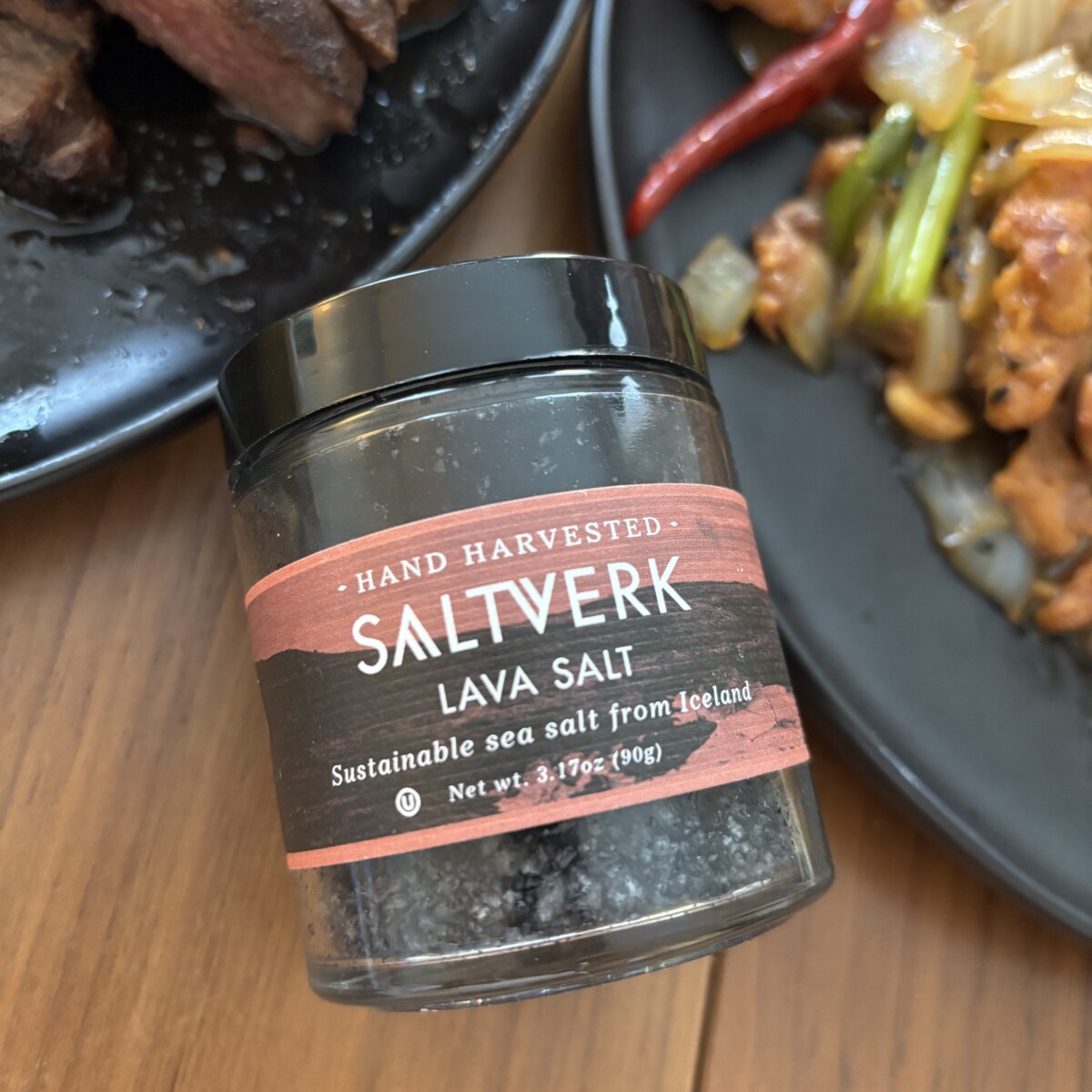 Jar of Saltverk Lava Salt from Iceland on a wooden table next to plates of grilled steak and stir-fried pork