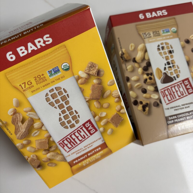 Two flavors of Perfect Bar refrigerated protein bars — Peanut Butter and Dark Chocolate Chip Peanut Butter — on a kitchen counter next to a Costco box