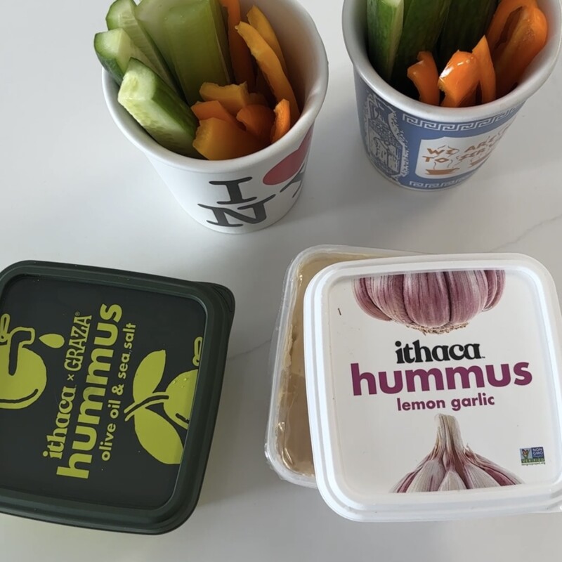 Five store-bought hummus containers including Ithaca x Graza, Ithaca Lemon Garlic, Cedar's Organic, Hope Foods, and Little Sesame arranged on a kitchen counter with vegetables and pita